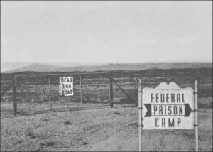 Photo shows a desert landscape with signs reading "Federal Prison Camp" and "Dead End Road"