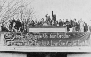 Photo shows a smiling, waving group of 20 people above a banner reading, "Hey people, smile on your brother; let me see you get together and love one another right now."